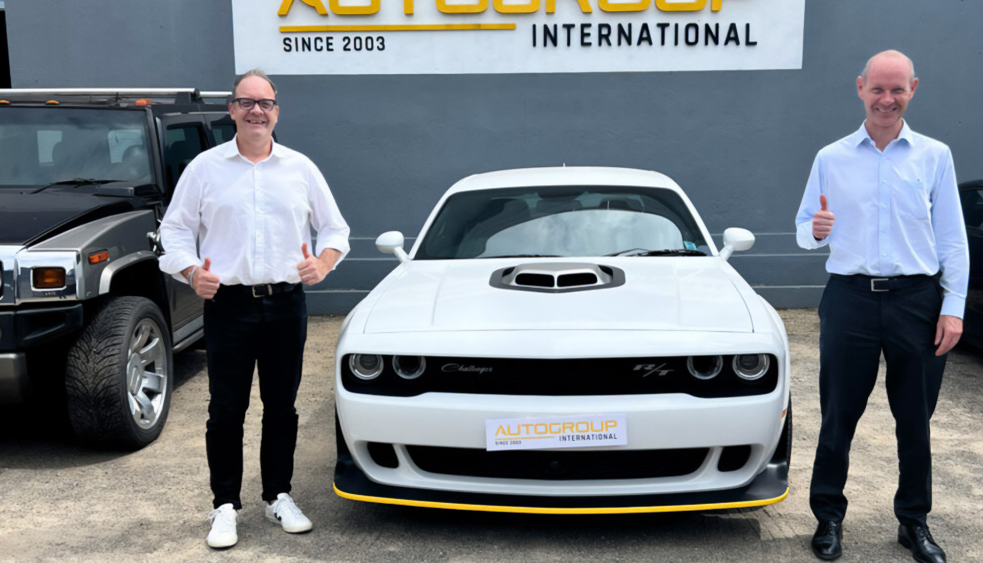 Two men posing with white Dodge Challenger at dealership.